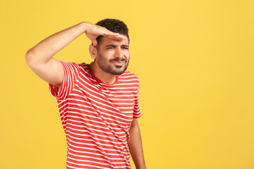 Fototapeta premium Nosy attentive man with beard in red striped t-shirt looking far away holding hand near forehead, watching at distance, searching for something. Indoor studio shot isolated on yellow background
