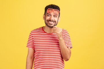 Excited positive man with beard in striped t-shirt holding party mask on stick and looking at camera with toothy smile, having fun. Indoor studio shot isolated on yellow background
