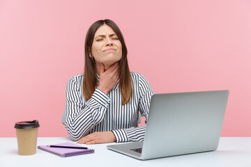 Sad depressed business woman touching neck feeling pain while swallowing, suffering thyroid inflammation, sore throat, sitting at workplace with laptop. Indoor studio shot isolated on pink background