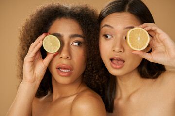 Portrait of two funny mixed race young women looking surprised, holding lemon and lime cut in half while posing together isolated over beige background