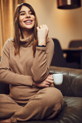 A pregnant woman sitting on a sofa at home with legs crossed and having a coffee break.