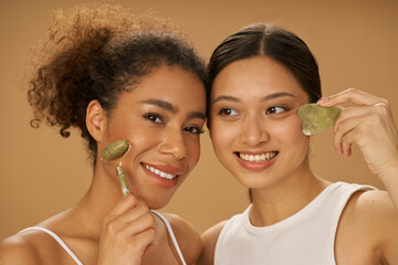 Portrait of lovely young women smiling while using jade roller and facial gua sha, posing together...