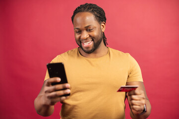 Cheerful African-American guy is using a smartphone and a credit card for shopping online, black man is ordering food online, male paying for long-awaited purchase on the mobile phone, isolated on red