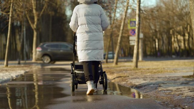 Mother with a baby carriage walking down the street woman pushing baby pram on path in town, winter or early spring season. High quality 4k footage