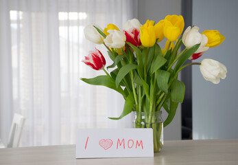 "I love Mom" card drawing with beautiful bouquet of colorful tulips in vase on table at home interior.  Congratulations and greetings for Mom on special day.