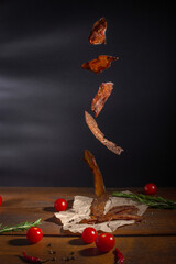 Food levitation. Flying meat jerky in front of dark background. Dried meat snacks, red cherry tomatoes, pepper, rosemary and peppercorns on craft pepper and wooden table. - obrazy, fototapety, plakaty