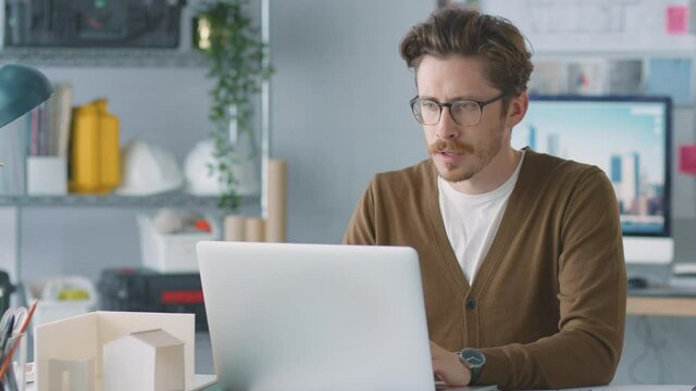 Camera tracks across frame as casually dressed architect makes video call from laptop at desk in office and shows drawings to client - shot in slow motion