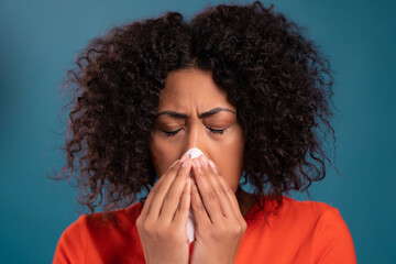 African woman sneezes into tissue. Isolated girl on blue studio background. Lady is sick, has a cold or allergic reaction. Coronavirus, epidemic 2021, illness concept