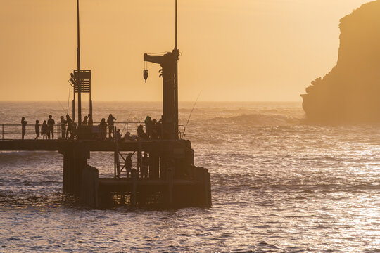 Fishermen and sightseers on a jetty in front of a golden sky