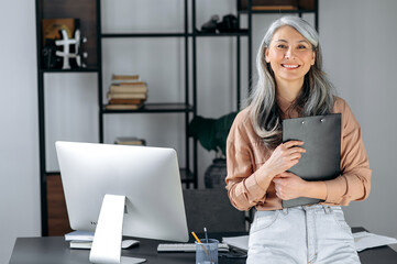 Portrait of a successful middle aged gray-haired asian business woman standing in modern office, dressed in stylish clothes, holding folder in hands, looking at camera, smiling friendly