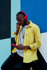 black guy with yellow shirt and red headphones and phone listening to music with his hand on his ears standing next to a colorful wall