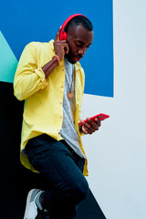 black guy with yellow shirt and red headphones and phone listening to music standing next to a colorful wall