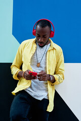 black guy with yellow shirt and red headphones and phone listening to music standing next to a colorful wall