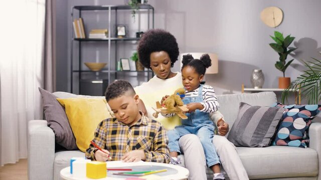 Beautiful African-American mother sits with two children at home, holding her little daughter in her arms, playing with toys while her son paints coloring book.