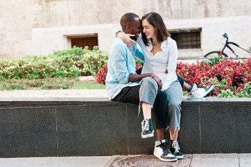 couple of young multiethnic guys sitting on a city bench hugging each other while talking, black guy and white girl in love