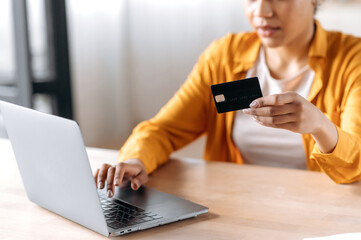 Online shopping and payment. Defocused African-American woman in casual clothes, sitting at the table, paying for purchases online, holds a card in her hand, entering credit card details into a laptop