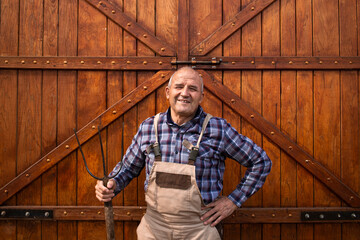 Portrait of smiling hardworking farmer holding pitchfork tool and standing by wooden barn or food granary doors at domestic animals farm.