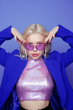 A girl in a blue jacket on a purple background in makeup in the style of the 70s, 80s, 90s, long silver earrings and purple glasses and a pink jacket. A girl in a shining jacket. Fashion style.
