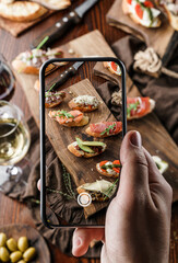 Woman's hands taking photo of set bruschetta with prosciutto, salmon, tomatoes, mozzarella, parmesan cheese, avocado on wooden background by smartphone device