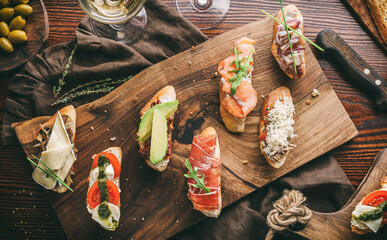 Fresh bruschetta with prosciutto, salmon, tomatoes, mozzarella, parmesan cheese, avocado and basil on rustic wooden background with glass of wine. Sandwiches for snack, top view, toning