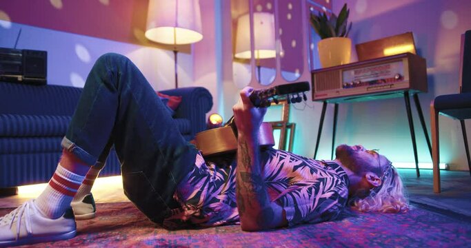 Young hipster in stylish shirt lies alone on floor singing and playing the guitar at home retro party in disco light.