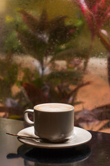 Closeup of a cup of coffee on a window with raindrops in winter weather