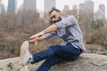 Young bearded man taking a break stretching body in the city park. Wearing formal shirt and jeans. Business office worker exercise concept