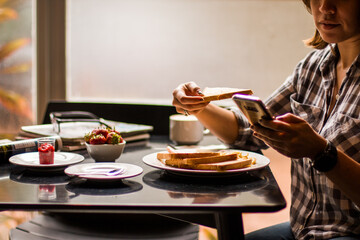 Arms of a woman holding a toast of bread and mobile phone with breakfast on a table served with eggs, ham, cheese and magazines