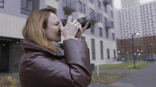 Side view of an adult caucasian woman taking pictures of a new multi storey residential houses. Action. Woman with a professional camera in winter weather outdoors.