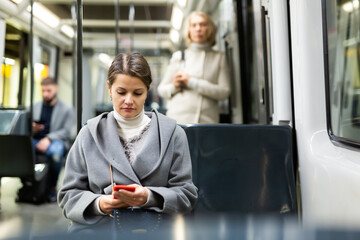 Young brunette browsing and typing messages on phone on way to work in modern streetcar