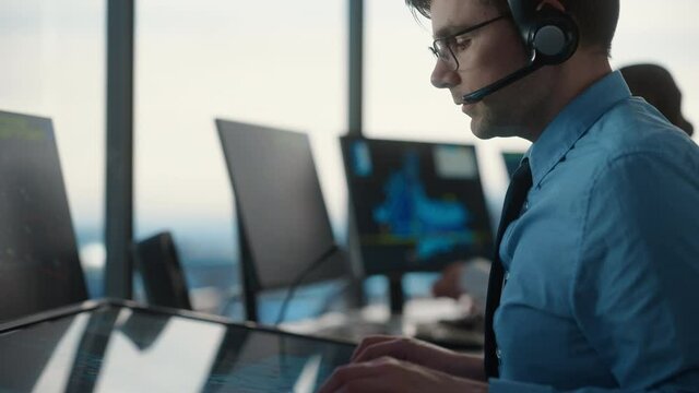 Male Air Traffic Controller with Headset Using Touchscreen Table Panel in Airport Tower. Office Room is Full of Computers with Navigation Screens, Airplane Departure and Arrival Data for the Team.