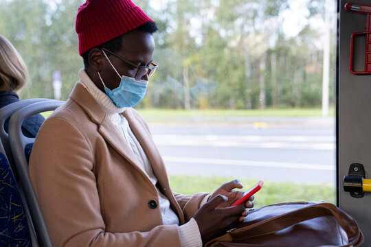 Stylish African American passenger man in beige coat, using cellphone, wear face mask to protect yourself from contact with flu virus, coronavirus Covid-19, traveling by public transportation. 
