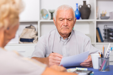 Upset senior man sitting at home table discussing with wife their bills