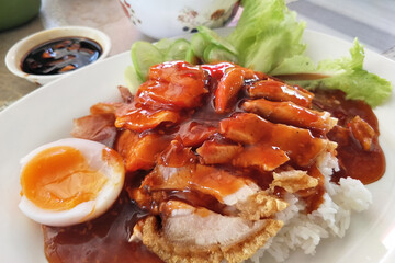 Rice with red barbecue pork, Crispy Pork, and Egg topped with sauce in a white dish. It is a delicious menu of Thai.