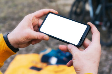 A man cyclist holds a mock-up of a smartphone with a white screen in his hands on the background of a bicycle in nature.