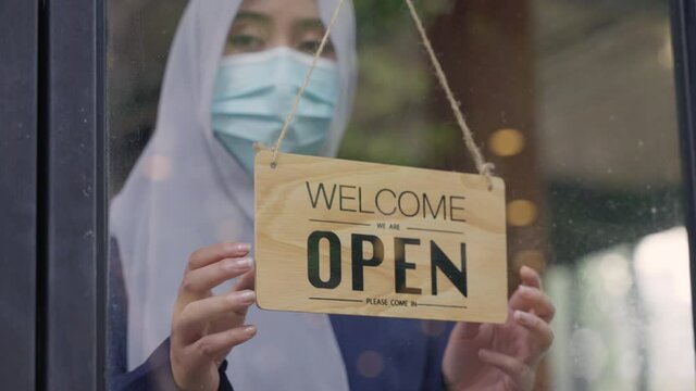 Young Asian Muslim woman wearing face mask turn entrance sign board to closed the coffee shop or restaurant due to coronavirus crisis. Female Muslim business owner changing sign from open to closed.