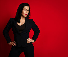 Self-confident slim woman in black pantsuit stands holding hands at waist, head turned looking at camera over red background with copy space. Fashion, style, trendy business look for female concept