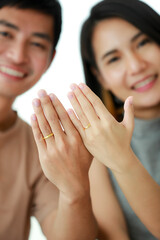 Portrait shot of cute smiling young Asian lover couple looking at the camera and showing a golden wedding rings that wore in their fingers. Selective focus at the rings isolated in white background