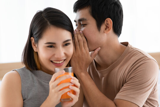 Portrait Shot Of Cute Smiling Young Asian Lover Couple Sitting On A Bed Together At Home In The Morning. Wife Holding And Drinking A Glass Of Orange Juice With Her Husband Whispering Her A Secret