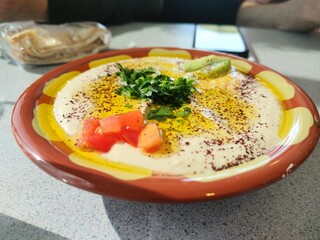 A bowl of fresh hummus with parsley, tomatoes, and olive oil sits on a restaurant table