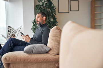 Caucasian adult man is sitting with smartpone in modern cozy office lounge zone