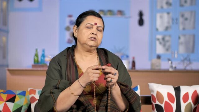 An aged Hindu woman chanting hymns / Mantras - religious faith concept. An Indian lady wearing a shawl / stole counting wooden beads while doing her daily prayers at home - spirituality 