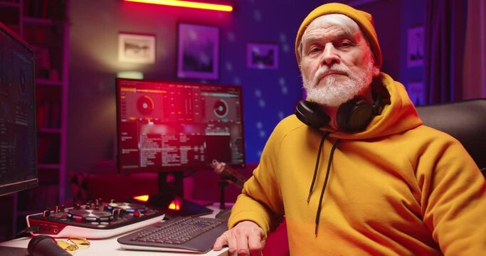 Close up portrait of stylish retired old handsome Caucasian male dj looking at camera and listening to song sitting in music recording home studio in evening. Mixing console creating music on computer