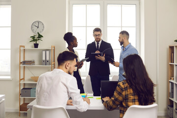 Multiethnic group of corporate employees and team leader working together with computers and paper document in busy open space. Office workflow, co-working, strartup business management