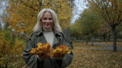 Closeup shot of happy elderly lady having fun with foliage in autumn day. 