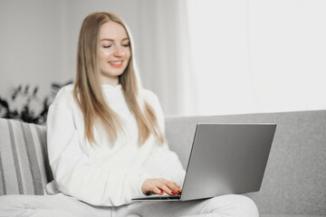 Young blonde woman sitting at home on the couch with laptop smiles and works distance in quarantine during the coronavirus pandemic
