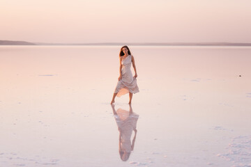 Young blonde woman in an evening airy pastel pink, powdery dress stands barefoot on white...