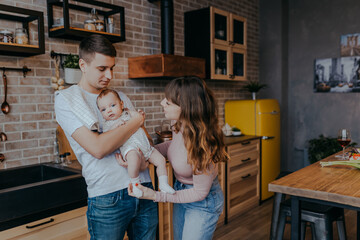 Fototapeta premium Young father holding baby boy in arms. Daddy and son together at home. Happy family in the kitchen. Mother looks at baby.
