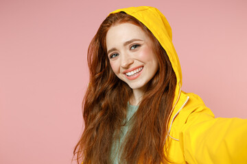 Redhead young smiling woman in yellow waterproof raincoat outerwear do selfie shot mobile phone isolated on pastel pink background studio portrait Outdoors lifestyle wet fall weather season concept