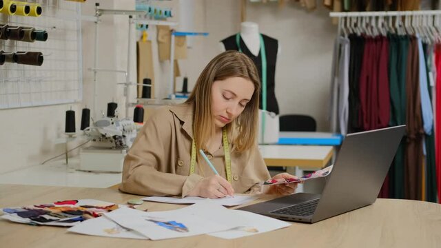 Close up of female designer is using her laptop computer working at the design studio. Tailor is looking at fabric samples and making notes. Clothing design start-up concept. Small business